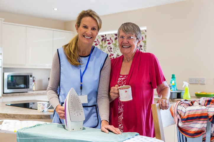 Care worker making a home visit. Female carer is ironing in the kitchen to help an elderly woman. They are both looking at the camera smiling.