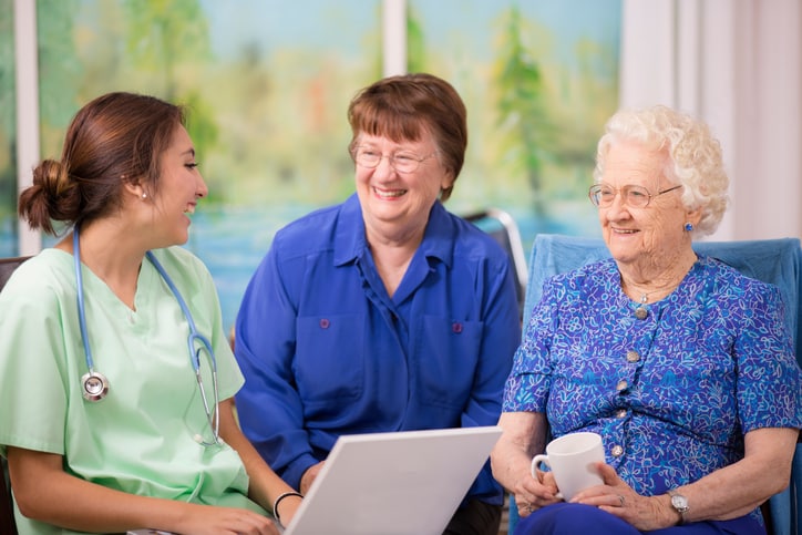 Female nurse conducts family consultation, takes notes on laptop. Elderly patient and her daughter meet with a caregiver after feeling a burnout